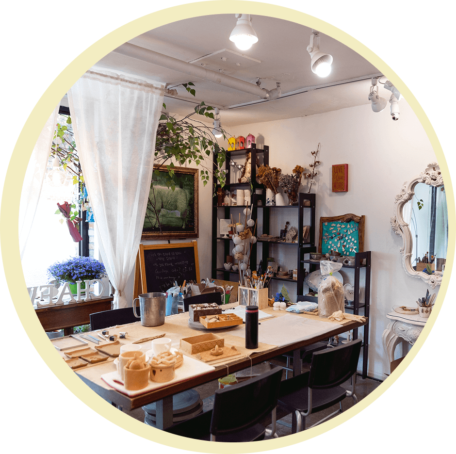 A cozy art studio with a wooden table, craft supplies, and decor pieces on shelves. A painting and mirror hang on the walls; a window with curtains lets in natural light.