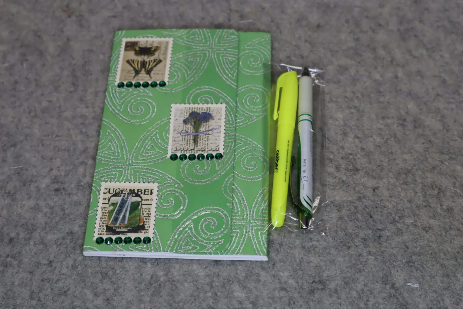 Green notebook with stamp designs and two pens on a gray surface.