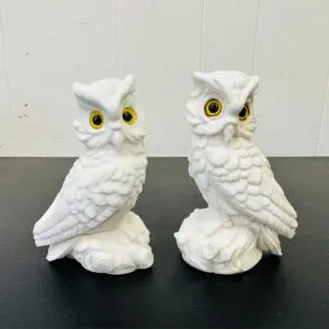 Two white ceramic owl figurines with yellow eyes on a black surface.
